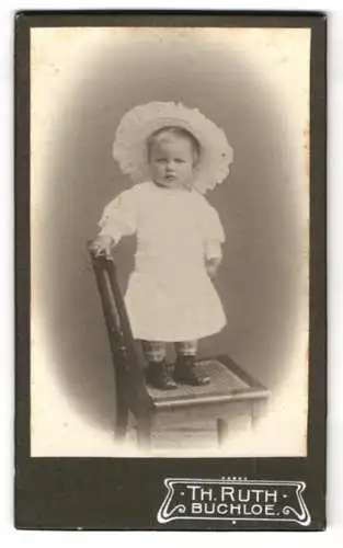 Fotografie Th. Ruth, Buchloe, Kleinkind im weissen Kleid mit grosskrempiger weisser Mütze