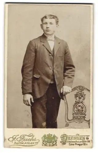 Fotografie J. Fuchs, Dresden, Waisenhaus Str. 16, Jugendlicher Knabe im Anzug mit einer Fliege, eine Hand auf dem Stuhl