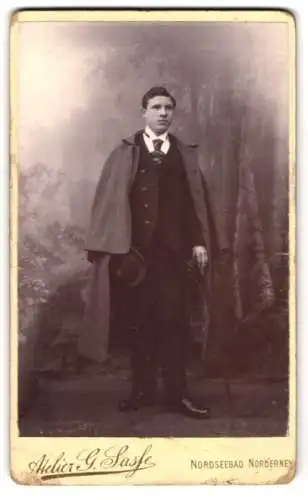 Fotografie Atelier G. Sasse, Norderney, Damenpfad 21, Junger Mann im Anzug mit übergeworfenem Mantel und Hut in der Hand