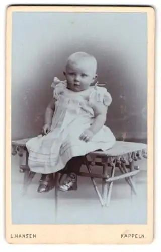 Fotografie L. Hansen, Kappeln, Blondes pausbäckiges Kleinkind im weissen Kleid, mit abwartendem Blick
