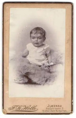 Fotografie F. G. Holle, Ilmenau, Südstrasse 3, Kleinkind im weissen Gewand, mit gespanntem Blick auf einem Pelz