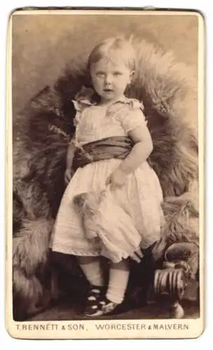 Fotografie T. Bennett & Son, Worcester, 8 Broad Street, Kleines Mädchen im weissen Kleid mit ihrer Puppe in der Hand