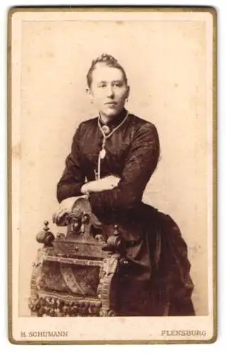 Fotografie H. Schumann, Flensburg, Holm 1, Junge Dame lehnt im schwarzen taillierten Kleid an einem Stuhl