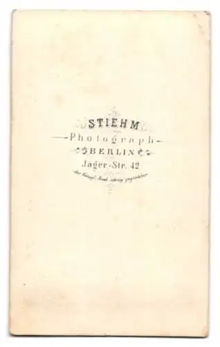 Fotografie Stiehm, Berlin, Jäger-Str. 42, junger Mann im dunklen Anzug mit Mantel und Fliege vor einer Studiokulisse