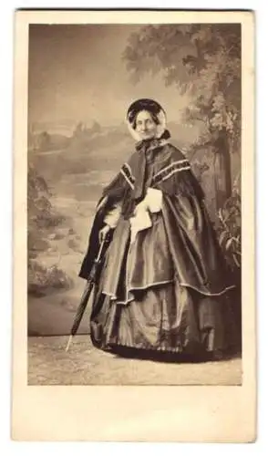 Fotografie Ferd. Liebsch, Hannover, ältere Dame im weiten dunklen Kleid mit Überwurd und Haube, Schirm, Studiokuliss