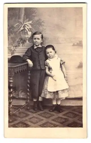 Fotografie unbekannter Fotograf und Ort, junger Knabe im Anzug mit seiner kleinen Schwester vor einer Studiokulisse