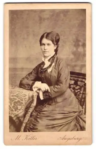 Fotografie M. Keller, Augsburg, Zeuggasse 226b, Junge Dame mit zurückgestecktem Haar und stoischem Blick an einem Tisch