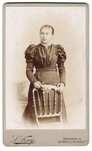 Fotografie L. Kny, Ebersbach i. S., Junge Dame mit lockigem Haar im schwarzen Kleid mit Hüftgürtel und Puffärmeln