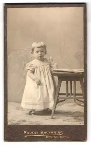 Fotografie Rudolf Zacharias, Regensburg, Kleinkind im weissen Gewand mit einer Kette, an einem Beistelltisch