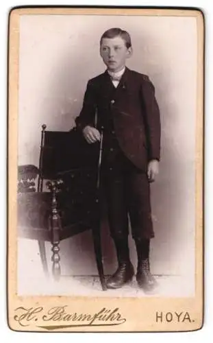 Fotografie H. Barmführ, Hoya, Bürgerlicher Knabe im Sonntagsstaat mit Seitenscheitel an einem Stuhl