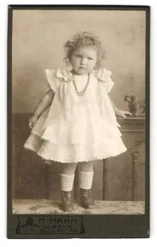 Fotografie H. Hahn, Lübeck, Gr. Burg-Str. 33, Kleines Mädchen mit lockigem Haar im weissen Kleid, mit zweifelndem Blick