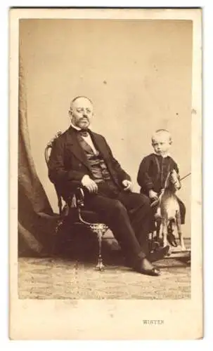 Fotografie M. L. Winter, Prag, Vater im Anzug mit Brille nebst seinem Sohn auf einem Schaukelpferd im Atelier