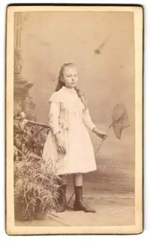 Fotografie A .Louvois, Bruxelles, hübsches junges Mädchen im weissen Kleid mit Schmetterlingsnetz vor einer Studiokulisse