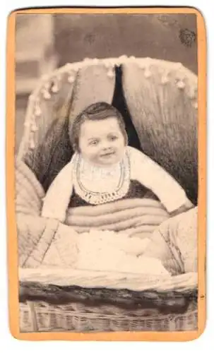 Fotografie unbekannter Fotograf und Ort, Kleines Kind zugedeckt im Bettchen sitzend, mit einem Latz
