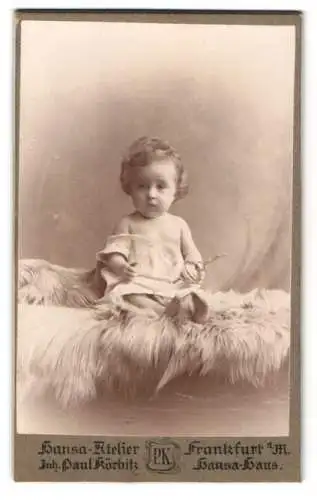 Fotografie Hansa-Atelier, Frankfurt a. M., Hansa-Haus, Kleines Kind im weissen Gewand mit einer Rassel auf einem Pelz