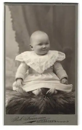 Fotografie Rob. Freimann, Heilbronn a. N., Fleinerstr. 24, Fast haarloses Kleinkind im weissen Kleidchen auf einem Pelz