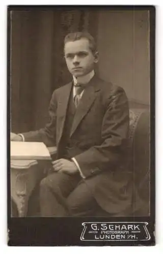Fotografie G. Schark, Lunden i. H., M. Schlüter im schwarzen Anzug mit offenem Buch am Tisch