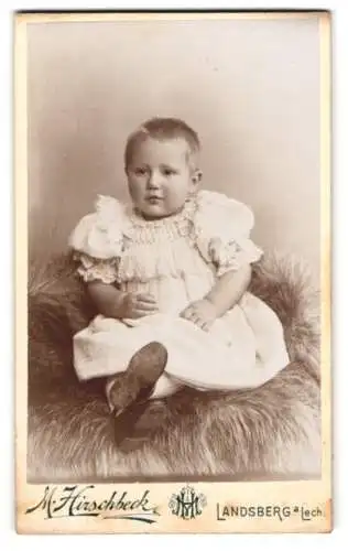 Fotografie M. Hirschbeck, Landsberg a. Lech, Niedliches Kleinkind im Kleid sitzt auf Fell