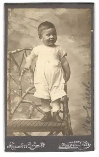 Fotografie Alexander Schmidt, Oberstein-Idar, Kleinkind Max Eissele in weisser Kleidung lächelnd auf einer Bank stehend