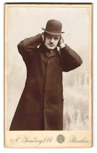 Fotografie A. Jandorf & Co., Berlin, junger Mann im Mantel mit Melone hält sich die Ohren zu, Schnappschuss