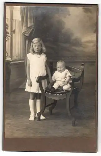 Fotografie unbekannter Fotograf und Ort, blondes Mädchen im weissen Kleid nebst Baby auf Sessel sitzend