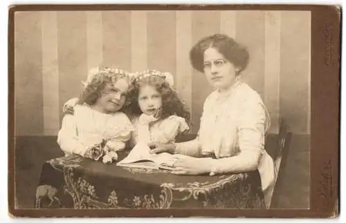 Fotografie Franz Zehetner, Mauerkirchen /Ob.-Oesterr., Junge Frau im Kleid und zwei Mädchen mit Buch am Tisch