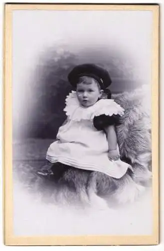 Fotografie R. Arndt, Aschersleben, Wilhelm-Strasse 1, Kleinkind mit Baskenmütze im Spitzenkleidchen auf einem Fell