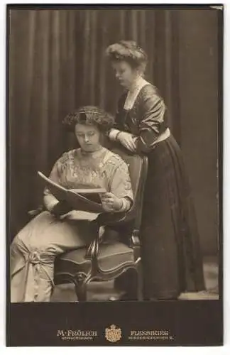 Fotografie M. Fröhlich, Flensburg, Norderhofenden 9, Zwei junge Damen in festlichen Kleidern, über ein Buch gebeugt