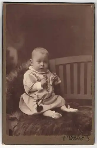 Fotografie J. Axer, Köszeg, Király Ut. 27, Baby im bestickten Kleidchen auf einem Fell