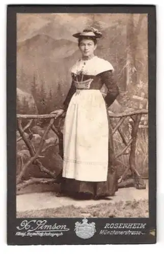 Fotografie X. Simson, Rosenheim, junge Frau in bayerischer Tracht mit Hut vor einer Studiokulisse