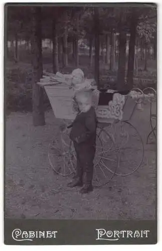 Fotografie unbekannter Fotograf und Ort, kleiner Junge steht neben Kinderwagen mit Geschwisterchen