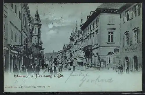 Mondschein-AK Freiburg i. Br., Strassenpartie mit Geschäften und Brunnen