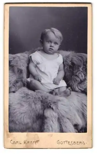 Fotografie Carl Krefft, Gottesberg, Blondes Kind im weissen Gewand auf einem Pelz, mit aufmerksamem Blick