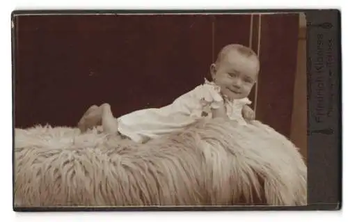 Fotografie Friedrich Kloerss, Rostock, Fröhliches Kleinkind im weissen Gewand auf einem Pelz