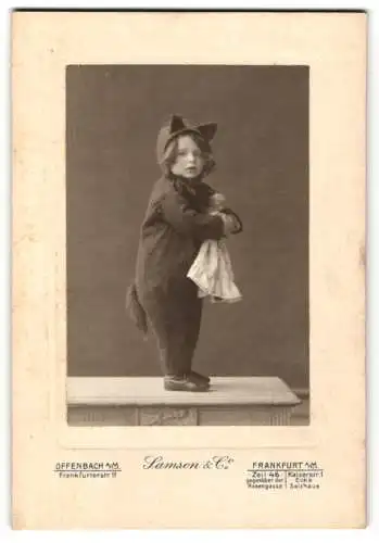 Fotografie Samson & Co., Offenbach a. M., niedliches kleines Kind im Katzenkostüm / Einteiler mit Puppe im Arm, Fasching