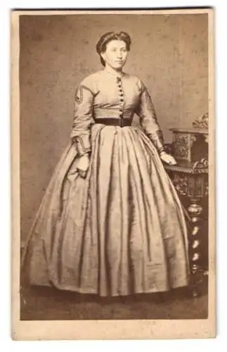 Fotografie C. Closter, Speyer, Dame im hellen Reifrockkleid stehend neben einem Sekretär