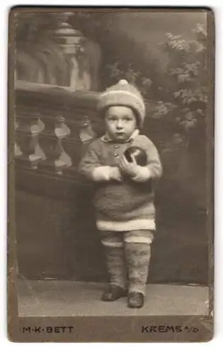 Fotografie M. K. Bett, Krems a. D., Dachsberggasse 5, Süsses Kind in Wollhemd und Mütze mit Ball im Arm