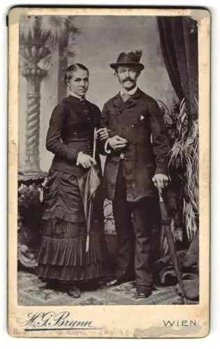 Fotografie H. G. Brunn, Wien, Margarethenstr. 22, Dame in tailleirtem Kleid nebst Ehemann und Regenschirmen