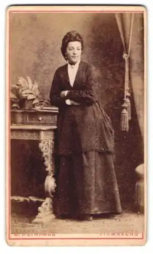 Fotografie G. Reinhard, Pirmasens, Elegante Dame in historischem Kleid neben verziertem Tisch