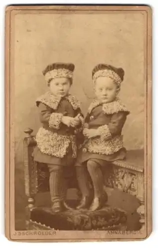 Fotografie J. Schroeder, Annaberg, Zwei kleine Kinder in winterlicher Kleidung auf dekorativem Möbelstück sitzend