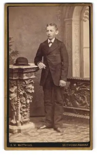 Fotografie E.W. Matthias, Seifhennersdorf, Portrait eines jungen Mannes in Anzug neben kunstvoll verziertem Podest