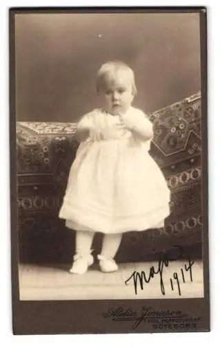 Fotografie Atelier Jonason, Göteborg, Kleinkind im weissen Kleid auf gemusterter Decke, 1914