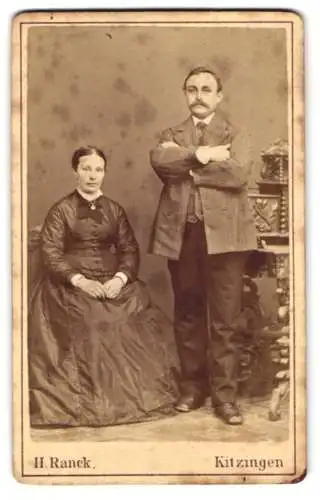 Fotografie H. Ranck, Kitzingen, Bahnhofstrasse, Sitzende Dame und stehender Herr mit verschränkten Armen