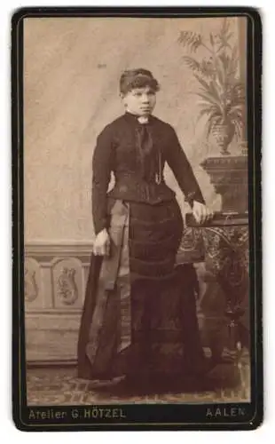 Fotografie G. Hötzel, Aalen, Porträt einer Frau in eleganter Garderobe neben einem Tisch mit Pflanze