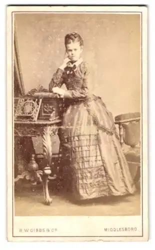 Fotografie R.W. Gibbs & Co., Middlesboro, Wilson St 20, Frau im viktorianischen Kleid an einem Tisch