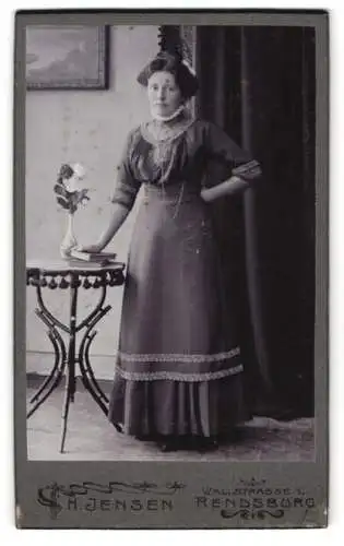 Fotografie H. Jensen, Rendsburg, Wallstrasse 1, Frau in elegantem Kleid neben Tisch mit Blumenvase