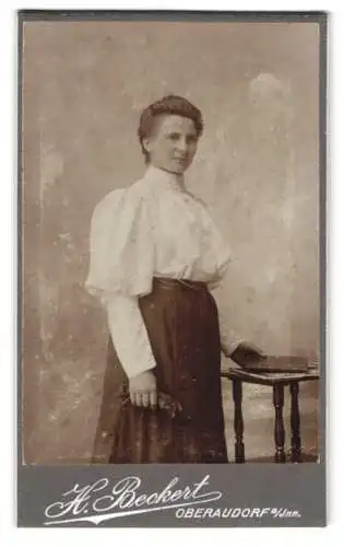 Fotografie Hans Beckert, Oberaudorf am Inn, Stehende Frau in Bluse und Rock neben Tisch