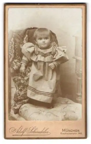 Fotografie Adolf Schindeler, München, Dachauerstr. 149, Kleinkind in langem Kleid auf gepolstertem Stuhl