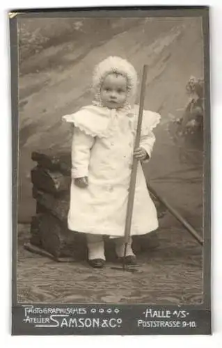 Fotografie Samson & Co., Halle a. S., Poststrasse 9-10, Kleines Kind im weissen Kleid mit Haube und kleiner Harke