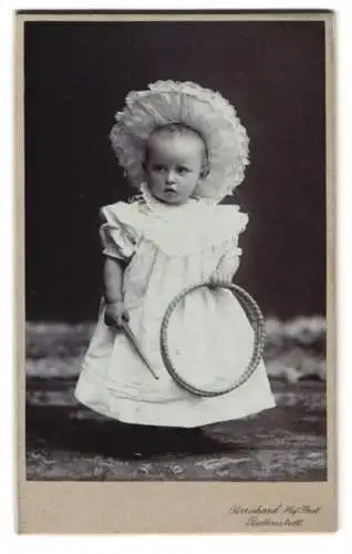 Fotografie Atelier Bernhard, Ballenstedt, Kleinkind im Kleid mit einer Haube und Spielzeug